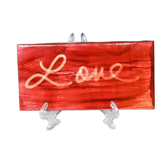 A 3X 6-inch ceramic tile with the word Love handwritten on a distressed looking background that has been painted with red alcohol ink.